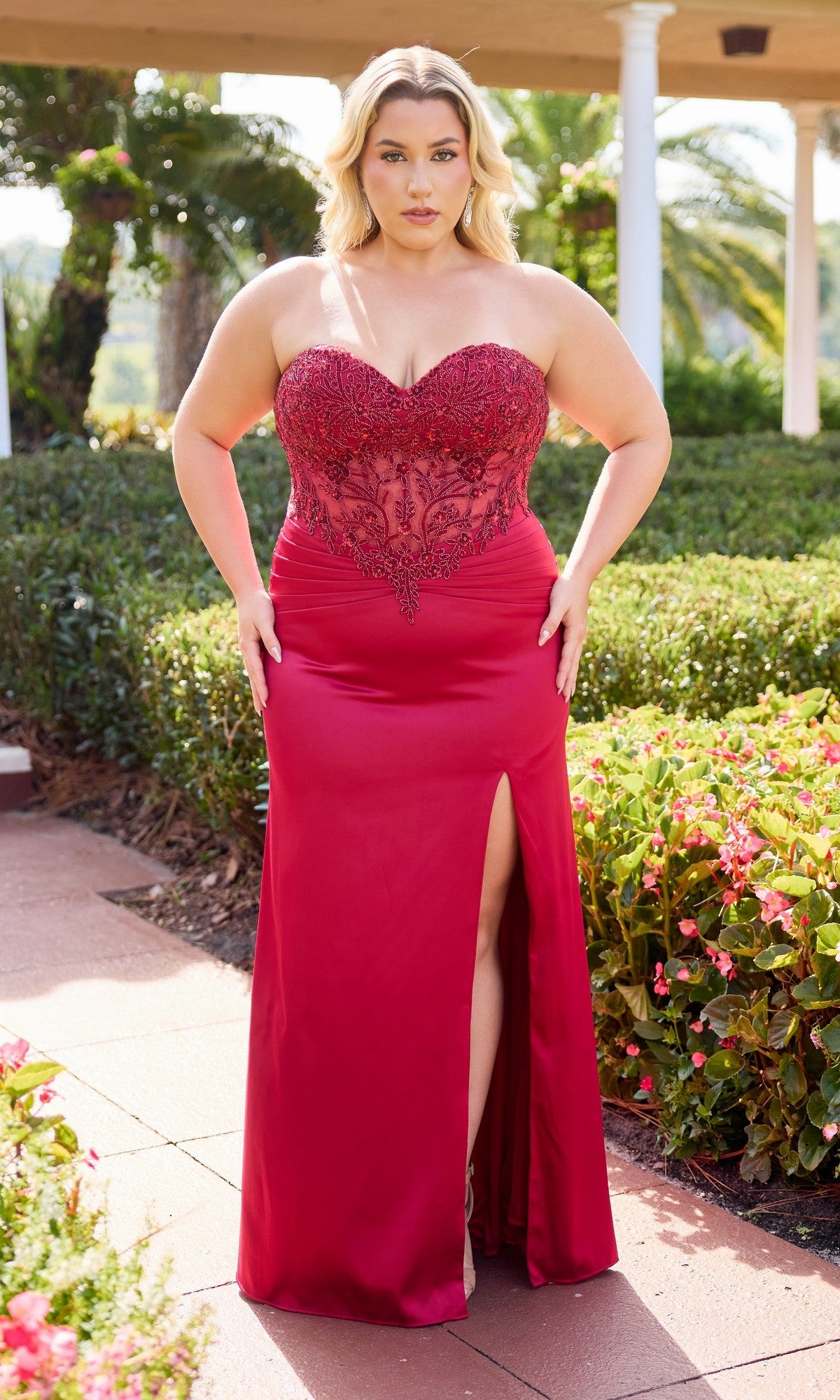 A woman stands outdoors in a red Primavera 14109 plus-size prom dress with a strapless sheer embroidered bodice, satin sheath skirt, and thigh-high slit, surrounded by vibrant greenery and flowers.